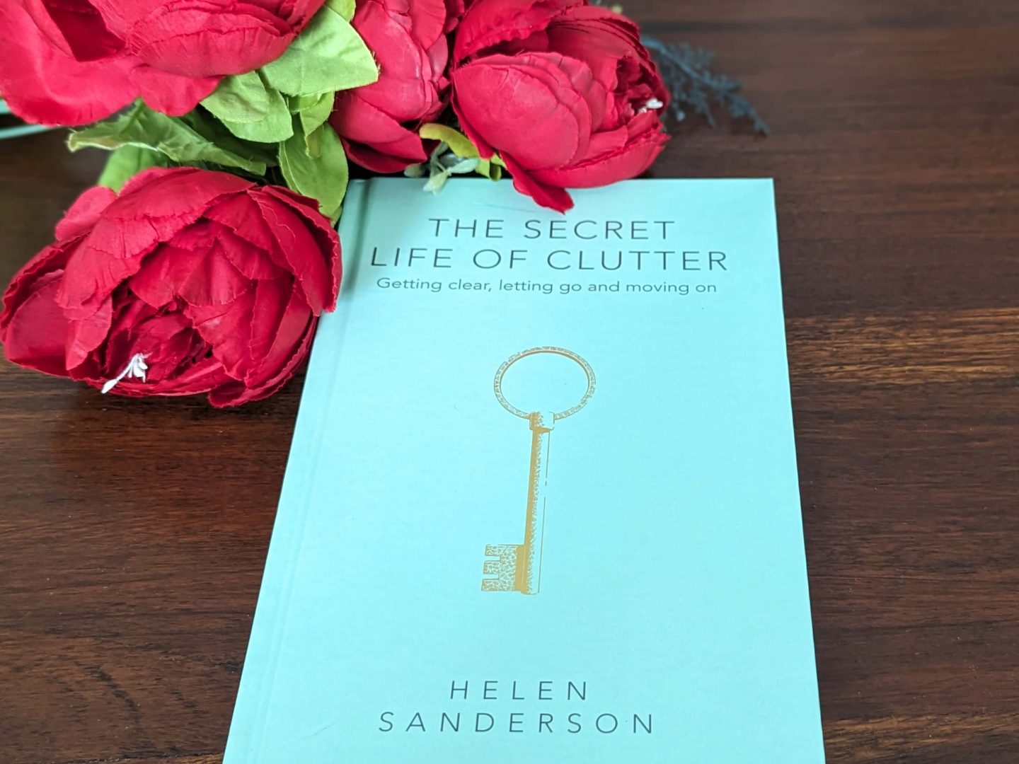 The secret life of clutter by Helen Sanderson - a book with a blue cover with a key on the front - displayed against a wooden background beside red flowers