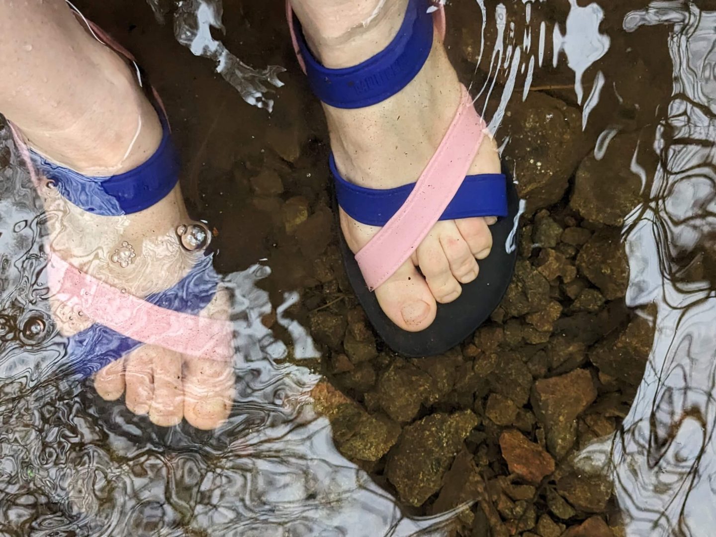 Female feet wearing pink and blue MOO CHUU sandals satnding in water