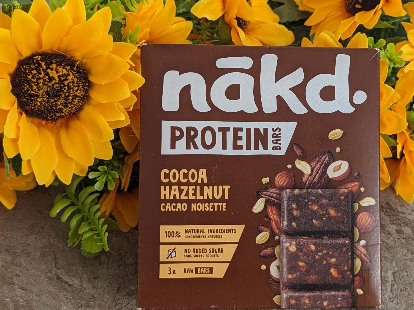 Pack of nakd vegan protein bars in cocoa and hazelenut flavour displayed against a bunch of sunflowers.