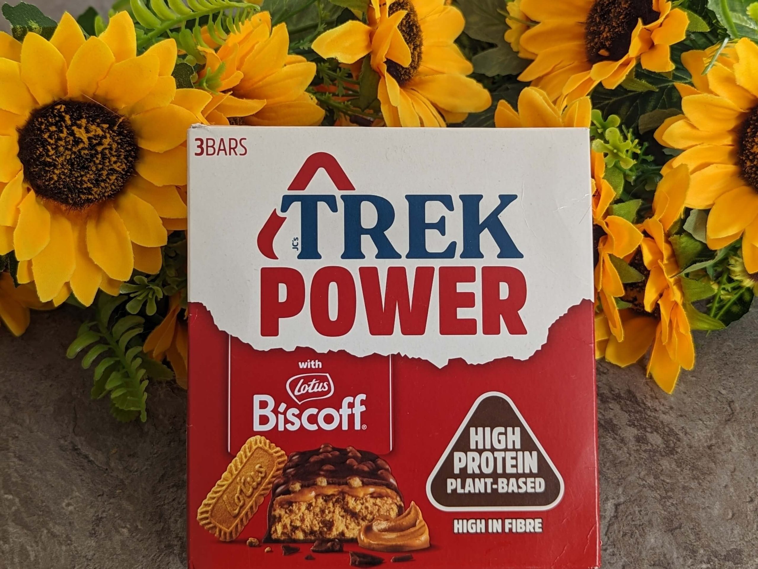 Box of Biscoff flavour vegan protein Trek Power bars displayed against a bunch of sunflowers.