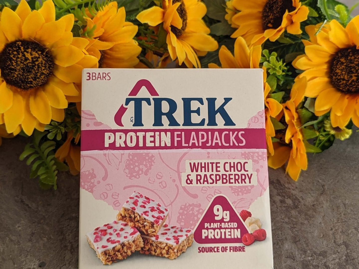 Box of Trek white choc & raspberry vegan protein flapjacks displayed against a bunch of sunflowers.