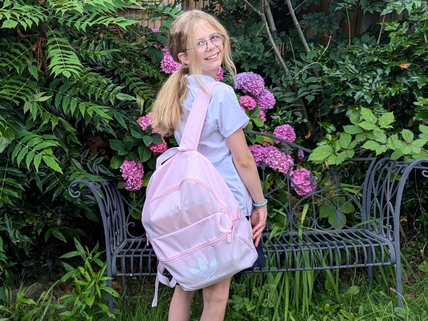 Libby wearing a white shirt and carrying a pink adidas backpack. She is standing in front of a garden bench and hydrangea. 