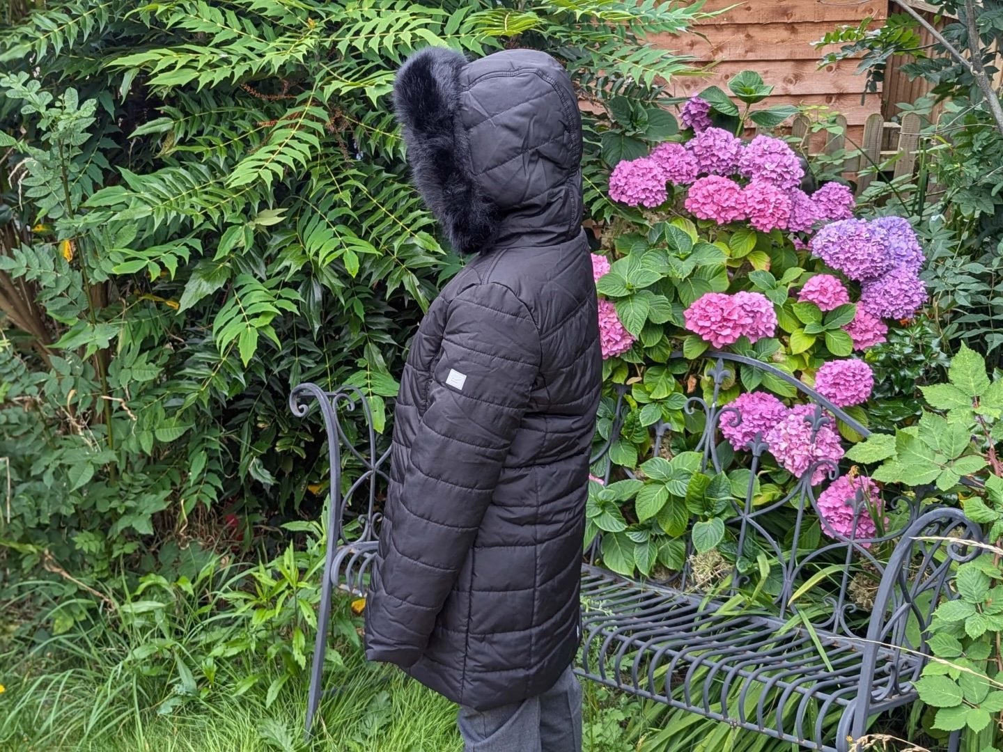 Lia facing sideways wearing a black regatta coat with the hood up so her face is not visible. She is standing in front of a garden bench and hydrangea