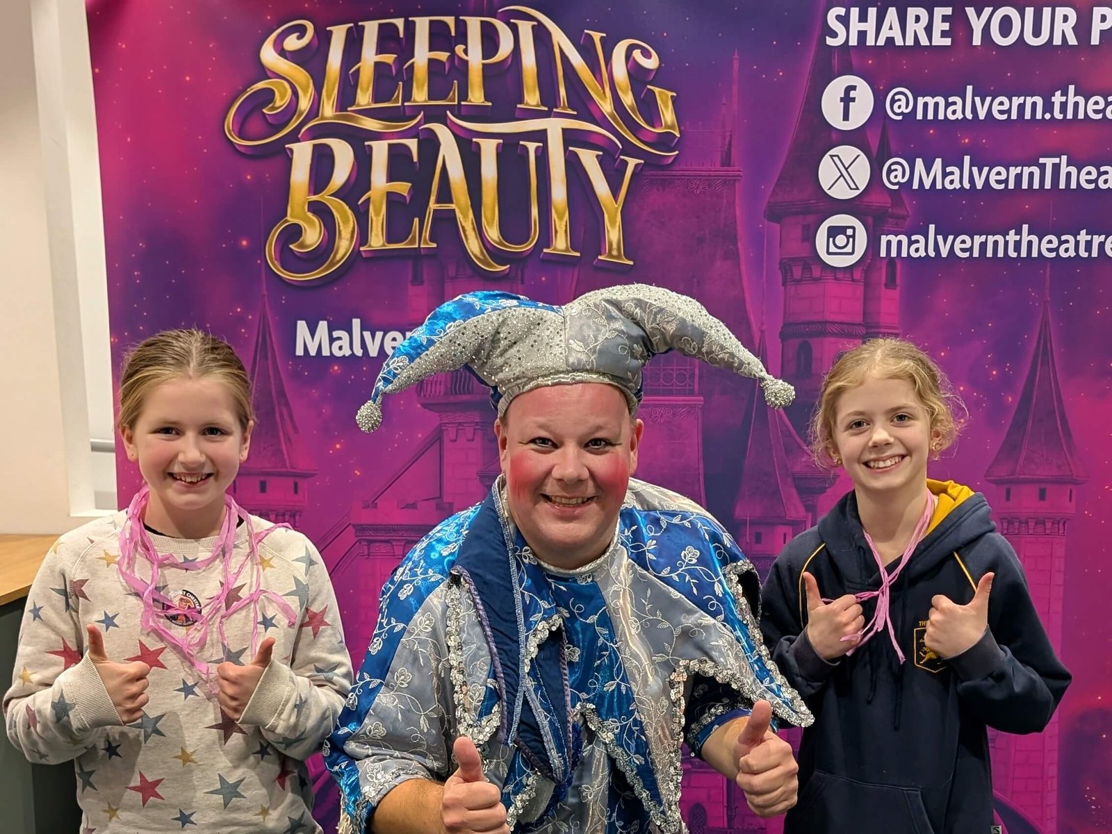 Mark James in front of a sign for Sleeping Beauty pantomime at Malvern with Two children all giving the thumbs up sign