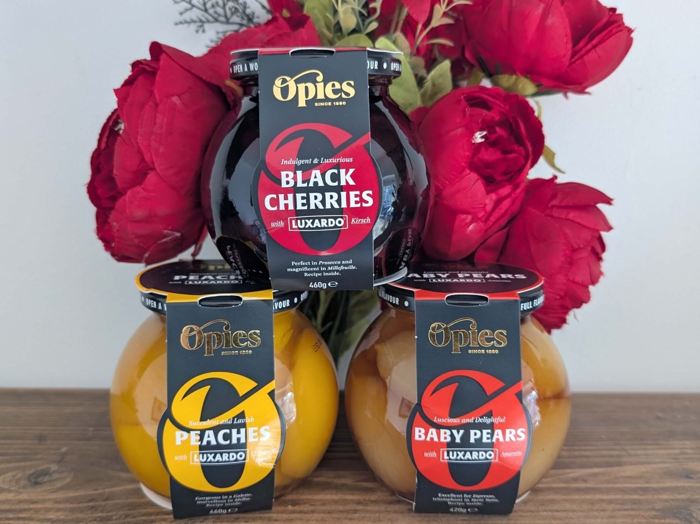 Three jars of Opies fruit in alcohol presented as a gift in front of red Christmas flowers on a wooden shelf