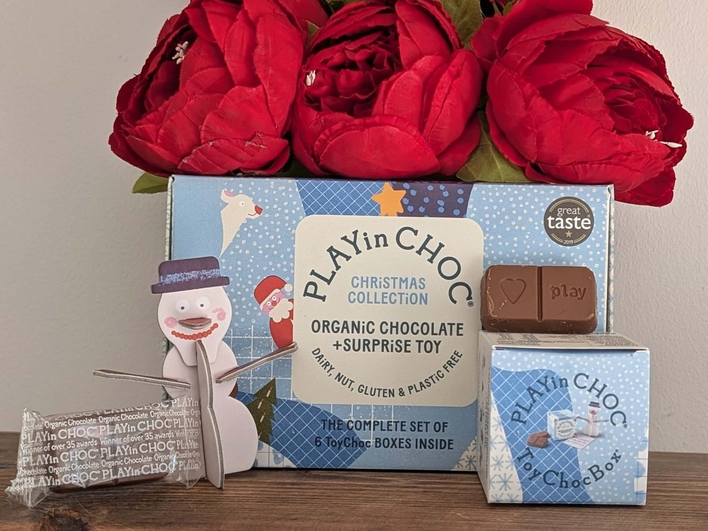 Play In Choc box with chocolate, smaller box and cardboard snowman displayed in front of red Christmas flowers on a wooden shelf
