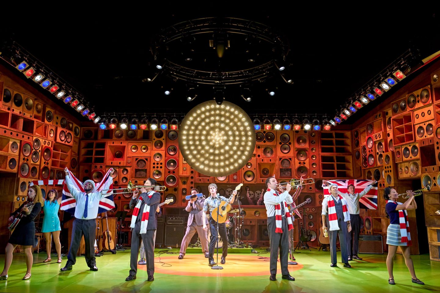 Band including trumpets and trombones on stage in Sunny Afternoon - The Kinks Musical with characters wearing red and white scarfs and waving a Union Jack flag