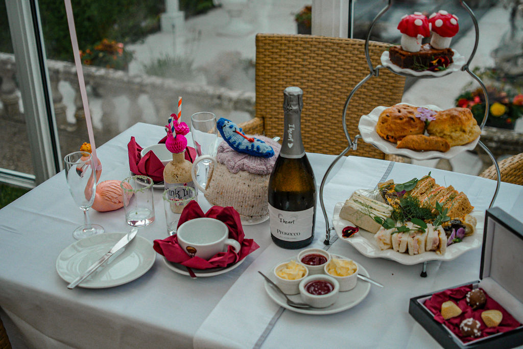 Alice in Wonderland themed afternoon tea at Ralph Court Gardens in Herefordshire, West Midlands image of a three tier afternoon tea with edible sweet toadstools on a chocolate brownie, scones, quiche and sandwiches and a themed tea pot with a bottle of prosecco and a flamingo on the table