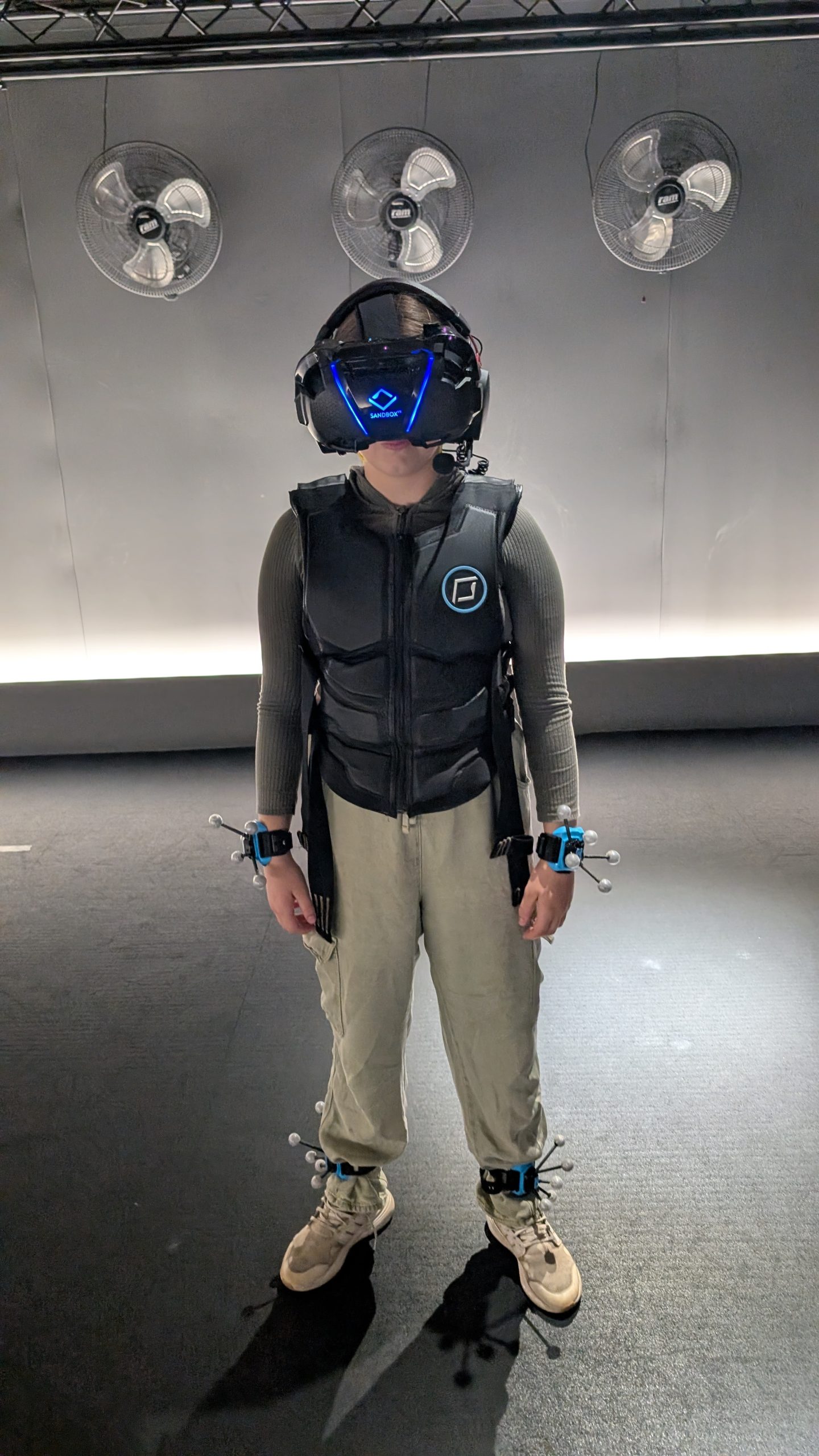Evelia standing in the VR room at Sandbox Birmingham wearing VR headset, black VR gilet and VR sensors on hands and ankles